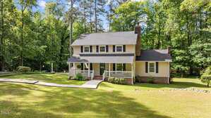Two-story yellow house with white trim and a covered front porch, set on a large grassy lawn with trees and shrubs in the background. A curved walkway leads from the driveway to the front steps.
