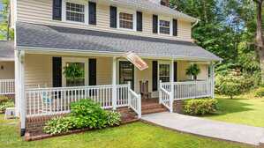 A beige two-story house with black shutters features a wide front porch with white railings, hanging ferns, a rocking chair, and an American flag. The porch overlooks a green lawn and is bordered by shrubs and trees.