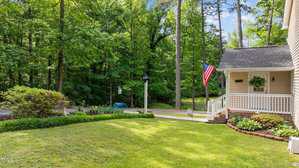 A neatly mowed front yard with a small porch, an American flag, and a hanging plant. The house is bordered by a wooded area with tall trees and a paved driveway leading to the street.