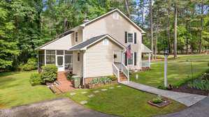 A two-story beige house with white trim, a screened porch, and an American flag on the front entry. The home is surrounded by grass, shrubs, trees, and a paved walkway leading to the entrance.