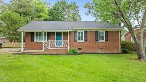 A charming one-story red brick house at 307 Pine Tree Road in Oxford, featuring white trim, black shutters, a small front porch with a white railing, and a blue front door, all set amid green grass and surrounded by trees.
