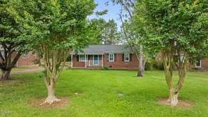 A single-story brick house with white trim, a small front porch, and black shutters is set back on a green lawn, framed by two leafy trees in the foreground. The sky is clear and blue.