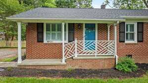 A brick house with a small covered porch, blue front door, white railings with a geometric pattern, black shutters, and green shrubs in front. The yard has grass and mulch with a few plants.