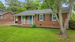 A one-story red brick house with black shutters, a covered front porch with white railing, and a blue front door. There is a green lawn, a tree in the yard, and another brick house partially visible on the left.