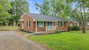 A single-story brick house with a dark shingled roof, blue front door, and white trim sits on a grassy lawn. A small porch with white railings is at the entrance. A gravel driveway runs along the side of the house.