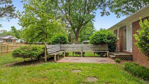 A wooden bench sits on a brick patio in a grassy backyard, bordered by bushes and trees. A red brick house with a white door is on the right, and a wooden fence encloses the yard.