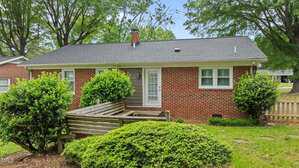 A single-story brick house with a gray shingle roof, white window frames, and a small wooden bench in the front yard. Green shrubs and trees surround the house, and there is a patch of grass in the foreground.