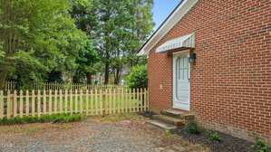 A brick house with a white door and striped awning, a small set of steps, and a gravel area in front. There is a light fixture by the door and a wooden picket fence with a gate enclosing a grassy yard with trees.