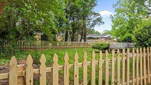 A backyard with green grass, surrounded by a light wood picket fence. Trees and bushes line the left side and background, with neighboring houses visible through the trees and fence.