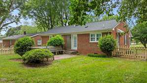 A single-story red brick house with a black shingled roof, white trim, and a small patio area with benches in a fenced backyard, surrounded by green grass, bushes, and mature trees.