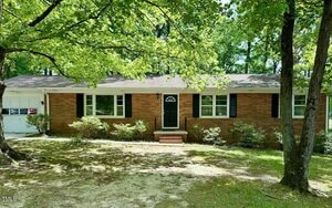 Charming single-story brick house at 1713 S Garnett Street, Henderson, with black shutters and a black front door. An attached white garage sits on the left. The yard is shaded by large trees, with minimal grass and some foundation shrubs.