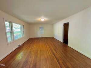 An empty room with hardwood floors, white walls, a ceiling light, three windows on the left wall, a single window on the far wall, and a doorway on the right leading to another space.