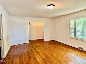 Empty room with hardwood floors, cream-colored walls, a ceiling light, a window letting in natural light, and an open doorway leading to another space. A floor vent is visible near the window.