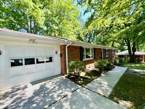 Single-story brick house with a white garage door, white trim, and a concrete walkway. The yard has shrubs and trees providing shade, and another house is visible in the background.