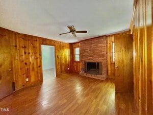 A wood-paneled living room with hardwood floors, a ceiling fan, a brick fireplace, two small windows, and two closed wooden doors. An open doorway leads to an adjacent room.