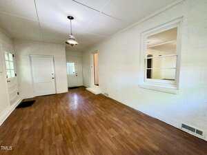 A rectangular room with white brick walls, wood flooring, a ceiling light fixture, two doors, a window opening to another room, and a small step leading to a hallway. The space is empty and well-lit.