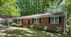 A single-story brick house with white-framed windows and black shutters, surrounded by trees and shrubs. There are steps leading to a black front door, and a driveway is visible to the left.