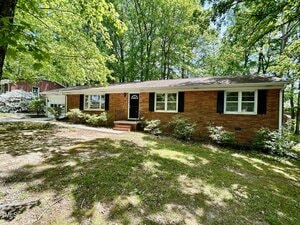 Single-story brick house with black shutters, white trim, and a dark front door, set among tall trees with a grassy and partially shaded front yard. Bushes line the front of the house.