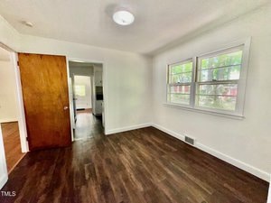 A small, empty room with white walls, dark wood flooring, a wooden door, and two windows letting in natural light. The room connects to a hallway and another space visible through the door.