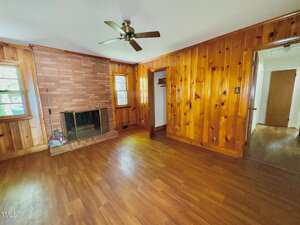 A living room with wood-paneled walls, a ceiling fan, a brick fireplace, two windows, an open closet, hardwood floors, and a doorway leading to another room. A broom is leaning by the fireplace.