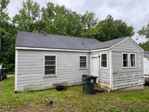 A small, single-story gray house with peeling paint and a black shingle roof, set on a patchy lawn with a trash bin near the back door. Trees are visible in the background under a cloudy sky.