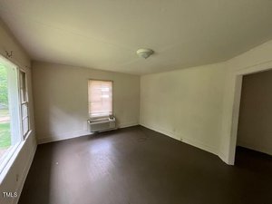 An empty room with brown floors, white walls, a ceiling light fixture, one window with a shade, and a window air conditioner below it. There is an open doorway on the right and a large window on the left wall.