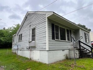A small, single-story gray house with wood siding, a white foundation, and black-framed windows. There are stairs leading to the front porch, an air conditioner in the window, and a satellite dish on the side. Grass surrounds the house.