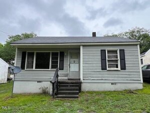 A small, single-story gray house with black shutters and a covered front porch. There are three wooden steps leading to the front door, and the yard has patchy grass. The sky is overcast.