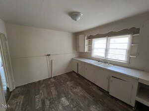 A small, empty kitchen with wood-look flooring, white cabinets, a sink under two windows, open shelves, and exposed plumbing connections; the walls and ceiling are painted white.