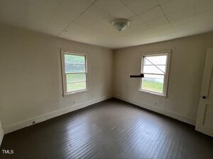 Empty room with two windows on adjacent walls, off-white walls and ceiling, a single ceiling light fixture, and a brown wooden floor. Daylight is coming through the windows.
