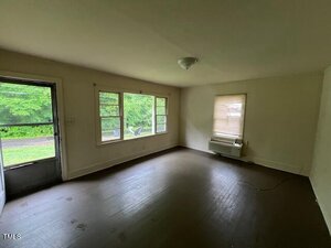 Empty room with brown flooring, white walls, three large windows, a single window with a blind, a ceiling light fixture, and a wall-mounted air conditioning unit beneath the smaller window. Door visible on the left.