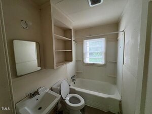 Small bathroom with off-white walls, a bathtub-shower combo, toilet, pedestal sink, wall mirror, built-in shelves, and a window with blinds letting in natural light.