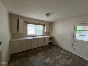 A small, empty kitchen with white cabinets, a sink under a window, open shelves, and a door with glass panes. The floor has a dark wood pattern, and the room is lit by a ceiling light.