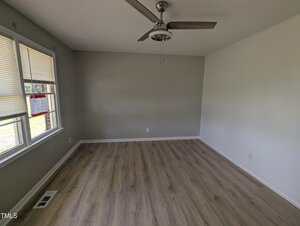 Empty room with light wood flooring, gray and white walls, a ceiling fan with two blades, and two windows with blinds partially open. A vent is visible on the floor near the windows.