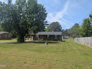 A one-story brick house with a covered front porch sits on a large grassy lot. A tree stands to the left of the house, and there is a stump or structure in the front yard. A wooden fence lines the right side.