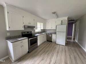 A kitchen with white cabinets, granite countertops, stainless steel stove and range hood, white fridge, dishwasher, and a window over the sink. The floor is light wood, and walls are light gray.