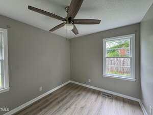 Small empty room with two windows, light gray walls, wood-patterned floor, a ceiling fan with wooden blades, and a view of a brick building and wooden fence outside.