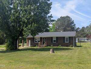 This charming single-story brick house at 1064 Abbott Road in Henderson features a covered front porch, black shutters, and an oval glass front door. A large tree and tree stump sit on the grassy lawn, with pine trees and houses in the background.