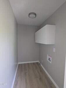 A small, narrow room with light gray walls, light wood flooring, a flush-mounted ceiling light, a white wall cabinet, and utility connections for laundry on the right wall.
