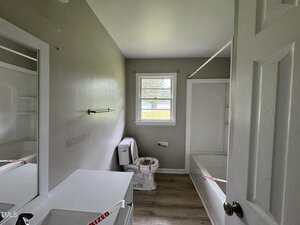 A small bathroom with gray walls, a toilet, bathtub with shower curtain rod, vanity with sink, towel bar, window, and wood-look flooring. The toilet and sink are secured with red "sanitized" tape.
