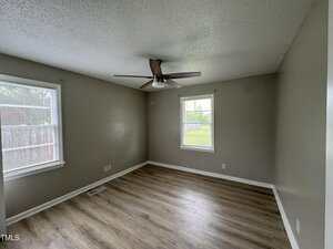 Empty room with light gray walls, light wood flooring, two windows with blinds, a ceiling fan with light, and a white baseboard along the walls. Natural light enters through the windows.