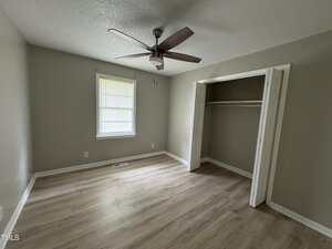 Empty room with light wood flooring, beige walls, a ceiling fan, a window with closed blinds, and an open closet with white doors. Natural light is coming in through the window.