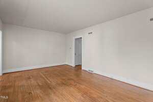 An empty room with white walls, a hardwood floor, a white door leading to another room, a white baseboard, and two air vents. The space appears clean with some marks on the floor and wall.