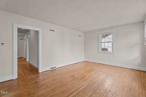 Empty room with light-colored walls, a wooden floor, a window with white trim on the right wall, and two doorways leading to other rooms. The space appears clean and well-lit by natural light.