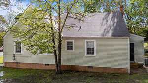 A small, light green house with white trim and a brick foundation, featuring two windows and a side door. A tree with green leaves partially obscures the view of the house. The yard has some patchy grass.