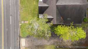 Aerial view of a house with a dark roof adjacent to a street with double yellow lines. The property has two trees with green foliage and a grass lawn next to a paved driveway.