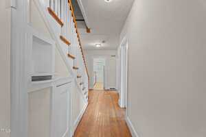 A hallway in a house with hardwood floors, beige walls, white trim, a staircase with a white railing on the left, and open doorways leading to other rooms; a small built-in shelf and cabinet are beneath the stairs.
