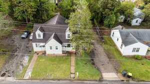 Aerial view of a light-colored house with a dark roof, front porch, and concrete walkway. A tree partially obscures the house. A driveway leads to a parked car. Adjacent is another house with garbage bins by the curb.