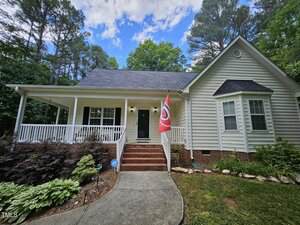 A light-colored single-story house with a porch, brick steps, and a red flag by the front entrance. The yard is landscaped with shrubs and plants, and tall trees surround the home under a partly cloudy sky.