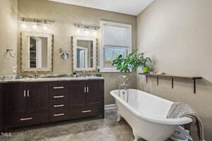 Bathroom with double sink vanity, two mirrors, overhead lights, a window, a freestanding clawfoot tub with a towel, and a shelf holding a potted plant and decor on beige walls.